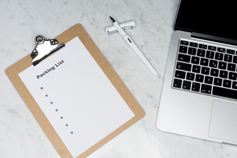 A top-down view of a workspace featuring a clipboard with a blank packing list attached, positioned on a white marble surface. Next to the clipboard is a white pen with a black tip, resting on the sur
