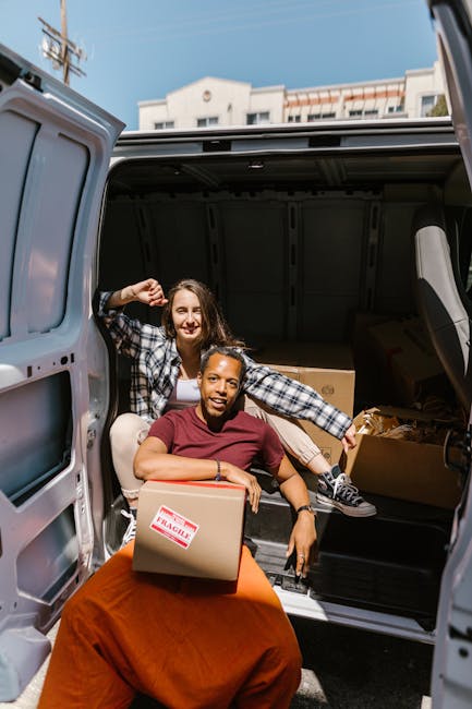 Two women are seen during the home relocation process, working together to load or unload boxes from the rear of a grey van parked outdoors under a clear sky. One woman, wearing a checkered shirt and 
