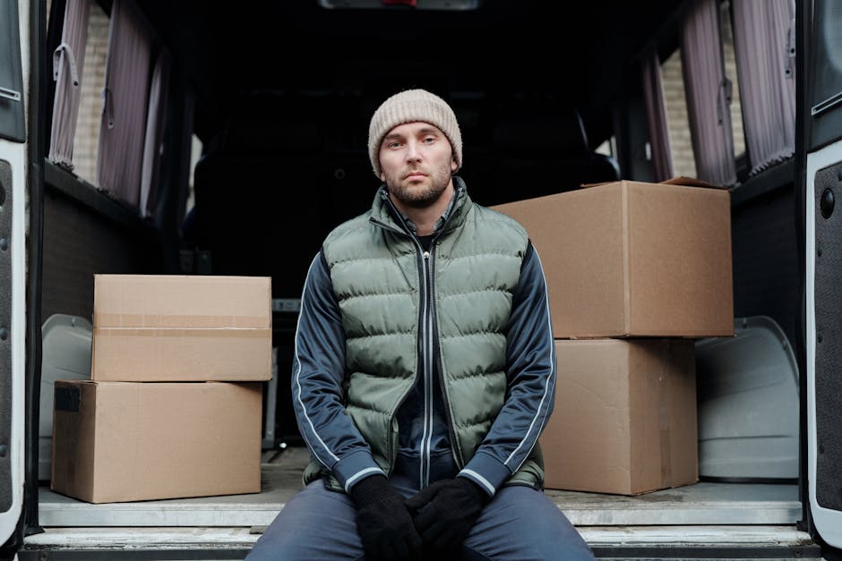 Two women are seen during the home relocation process, working together to load or unload boxes from the rear of a grey van parked outdoors under a clear sky. One woman, wearing a checkered shirt and 