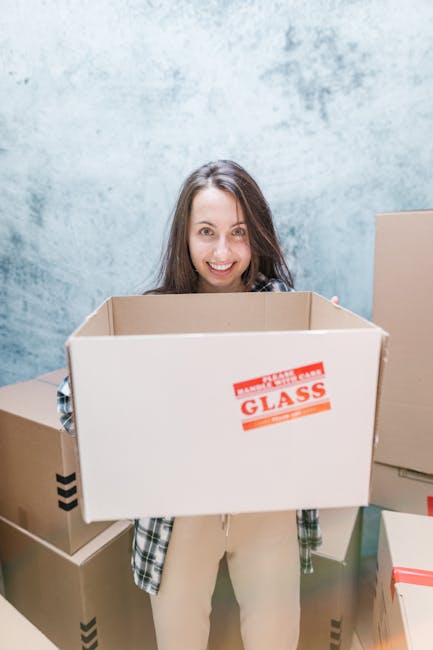 A young woman sitting cross-legged on a wooden floor surrounded by packed cardboard boxes with black printed labels and red 'Fragile' tape, inside a room with a light blue wall, as she plays with pack
